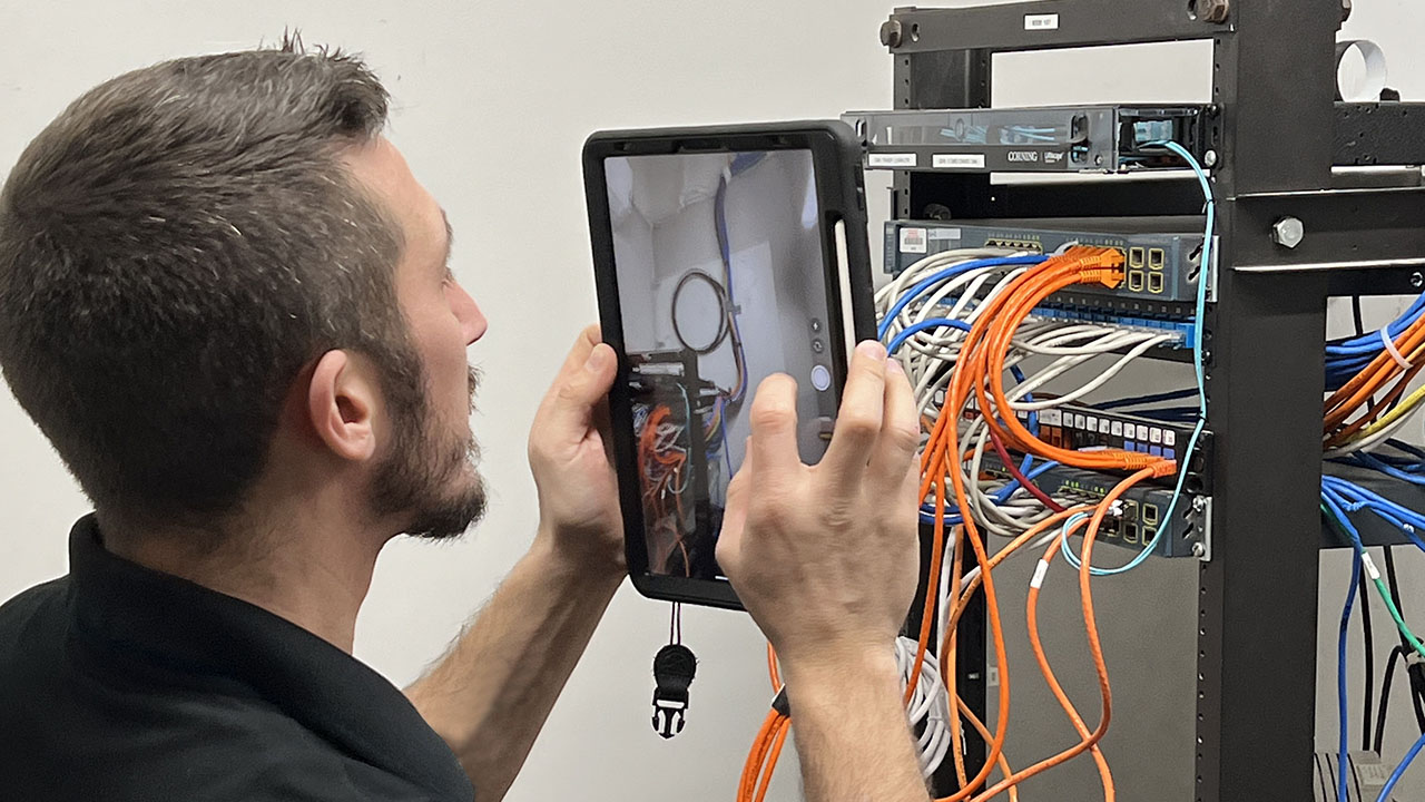 A Technology Specialist using a tablet to survey a Telecommunication Room IT equipment rack.